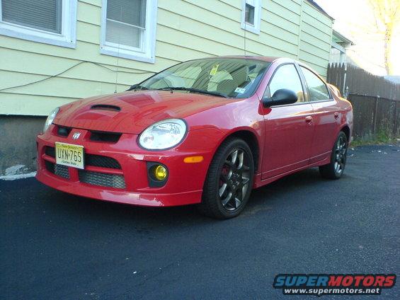 Red Dodge Neon SRT-4 with a hood scoop and yellow fog lights, parked on asphalt