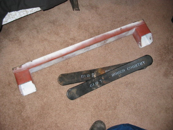 Two black metal bars with white lettering that says "ROUGH COUNTRY" and "GIO 7" sit on a carpet next to a red and white metal part