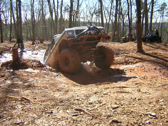 Off-road truck stuck in mud pit with trees around. Two ATVs are visible in the background