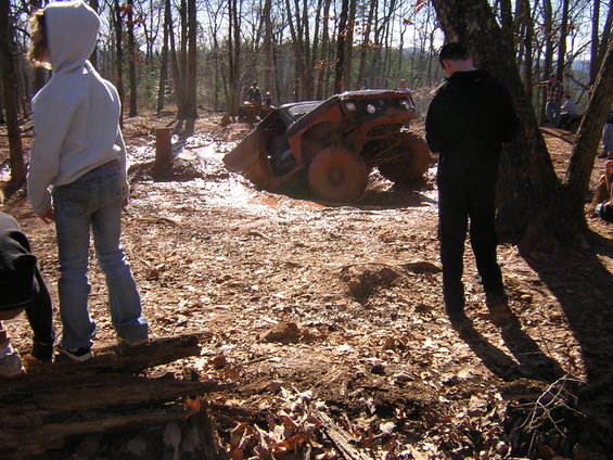 Orange off-road vehicle stuck in mud, front end angled towards camera, with people standing around in a wooded area