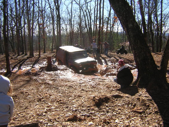 Off-road vehicle stuck in mud with people watching in a wooded area. Another ATV is visible in the background