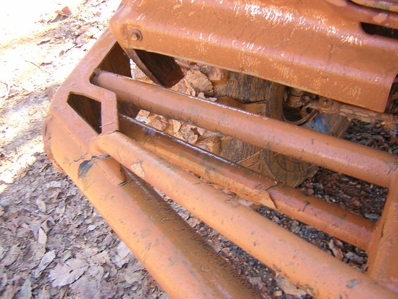 Close-up of a rusted orange grille with horizontal bars, partially obscuring a tire. Debris is visible on the ground