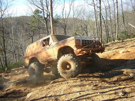 Mud-covered 1978 Ford Bronco climbing a steep, rocky trail with trees in the background