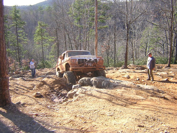 Muddy off-road truck climbing over rocks with two people watching