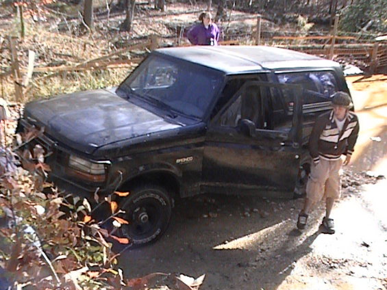 Black SUV, possibly a Ford Bronco, parked off-road with driver's door open. Two people are visible near the vehicle