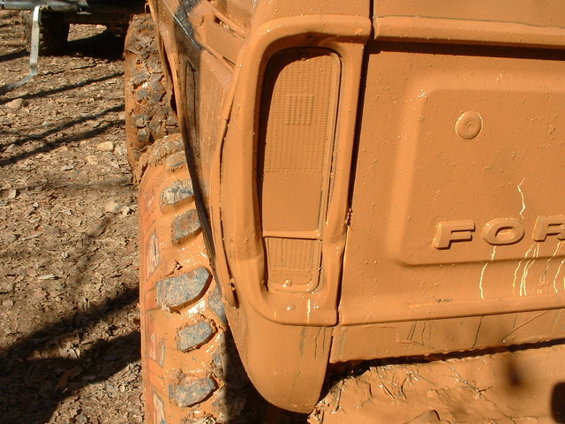 mud-covered truck rear quarter panel with taillight and 'FOR' emblem visible, next to a large off-road tire