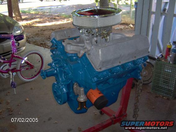 Blue V8 engine on red stand, chrome air cleaner, orange oil filter, next to bicycle and car