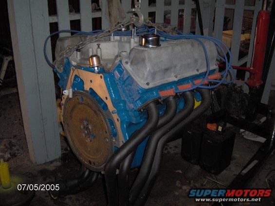 V8 engine block painted blue with silver valve covers and black headers, suspended by chains