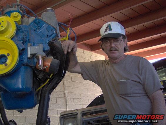 Man in a cap leans on a blue engine with yellow pulleys. Another vehicle is partially visible behind him