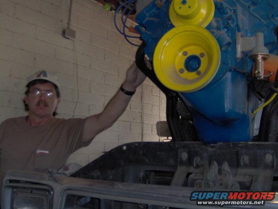 Man in tan shirt and cap works on a blue engine with yellow pulleys. Part of a vehicle's front end is visible below