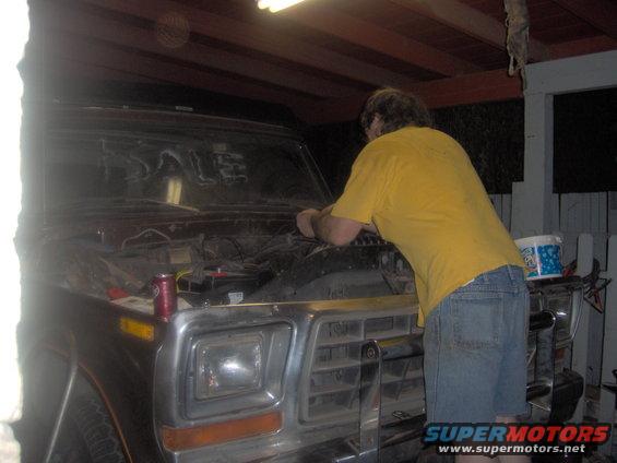 Person working on the engine of a 1978 Ford Bronco. Hood is open, showing engine components. A can of soda is on the fender