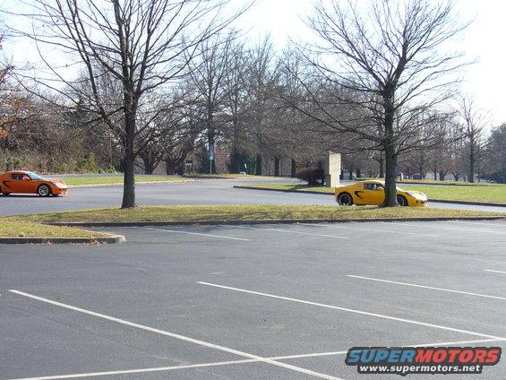 Orange sports car on left, yellow sports car parked near a tree on right. Parking lot scene