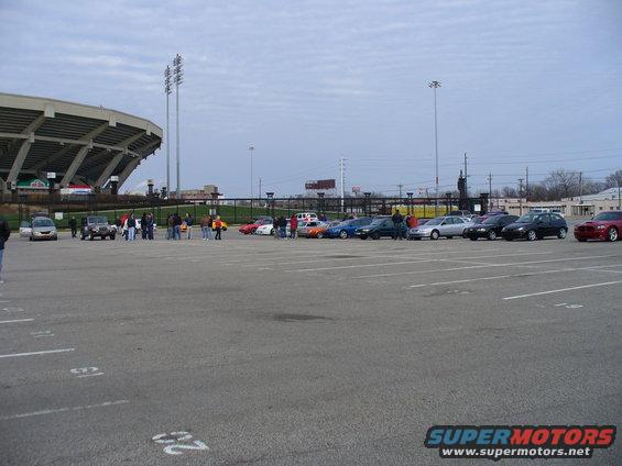 Parking lot scene with a stadium in the background and several cars parked in rows