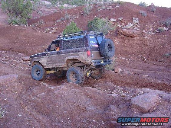 Muddy 1986 Ford Bronco II climbing a rocky trail with a spare tire on the back