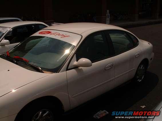 White sedan parked, side and front quarter view. Red 'Ford Racing' banner on windshield
