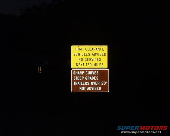 Yellow and brown road signs warning of high clearance, no services, sharp curves, and steep grades
