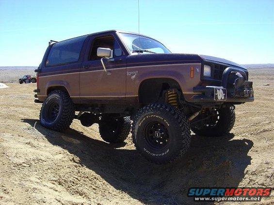 Brown Ford Bronco II with lifted suspension and off-road tires on a dirt hill. Another vehicle is visible in the distance
