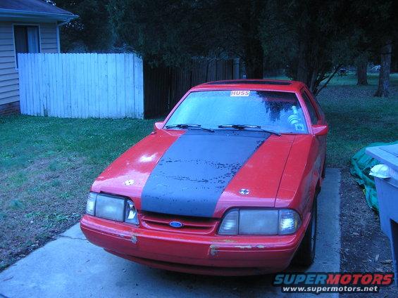 Red Ford Mustang, front view, with a black stripe on the hood and a 'HUSS' sticker on the windshield