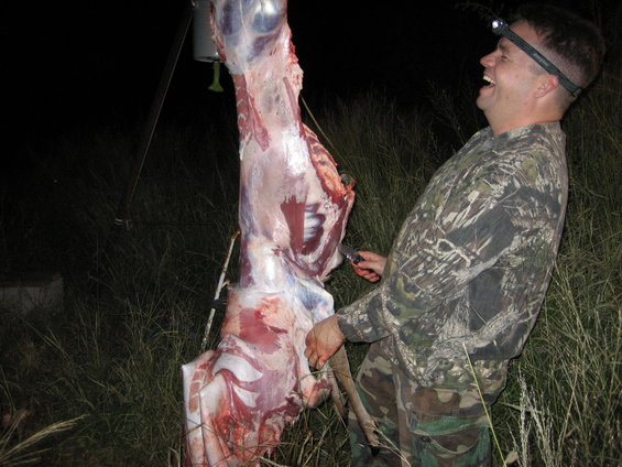 Man in camouflage laughing while butchering a large animal carcass hanging from a metal pole at night