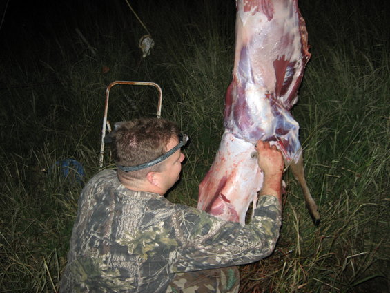 Man in camo with headlamp skinning a deer carcass hanging from a rope outdoors at night