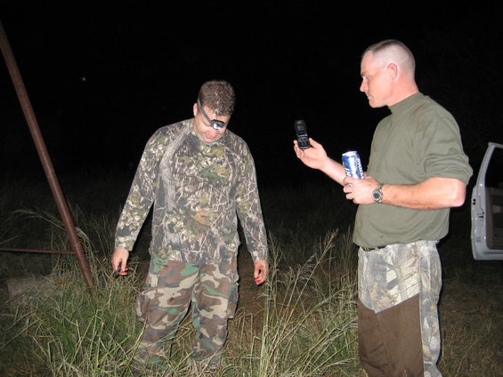 Man in camo shirt and pants with headlamp looks down. Another man in a green shirt and camo pants holds a flip phone and a can