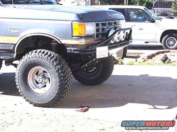 Front end of a lifted pickup truck with big tires and a brush guard. Another white SUV is parked behind it