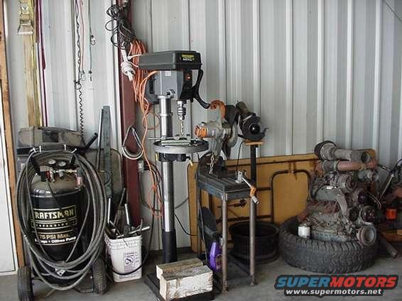 Workshop scene with a drill press, air compressor, and an engine block on a tire