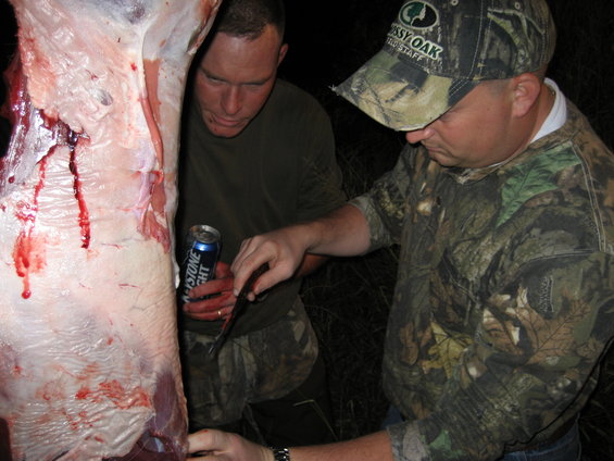 Hanging animal carcass with two men in camouflage clothing preparing to butcher it; one man holds a beer can