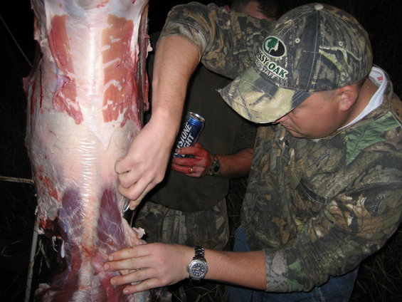 Guys in camo processing a deer carcass. One holds a beer