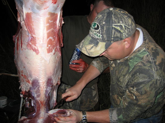 Deer carcass being processed by two men in camouflage clothing, one holding a can of Keystone Light