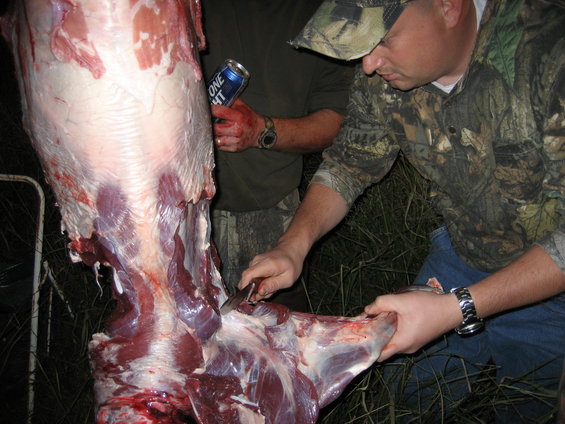 Guys in camo are butchering a big animal carcass. One is holding a beer