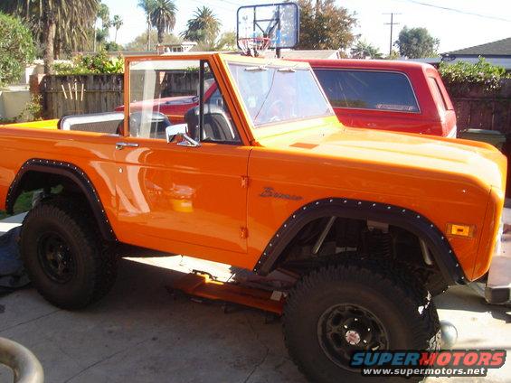 Orange 1977 Ford Bronco with black fender flares and a basketball hoop on the roof. Red SUV parked behind it