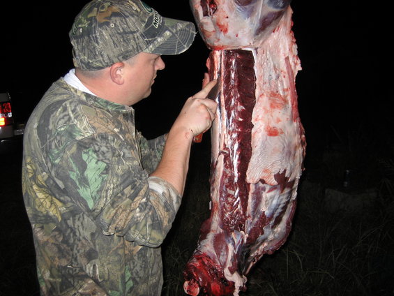 Man in camo cutting into a hanging deer carcass at night. A truck is visible in the background
