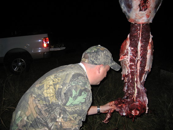 Man in camo looks at a deer carcass hanging near a white pickup truck at night