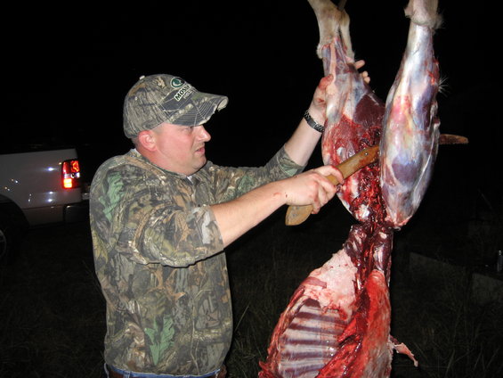 Man in camo hat and jacket butchers a deer carcass hanging from above. A white truck is visible in the background