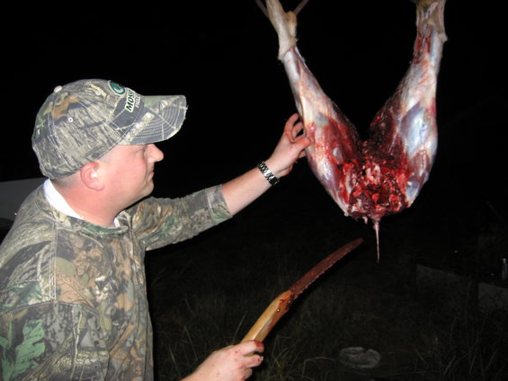 Man in camo holding a deer carcass by the hind legs. He has a saw in his other hand
