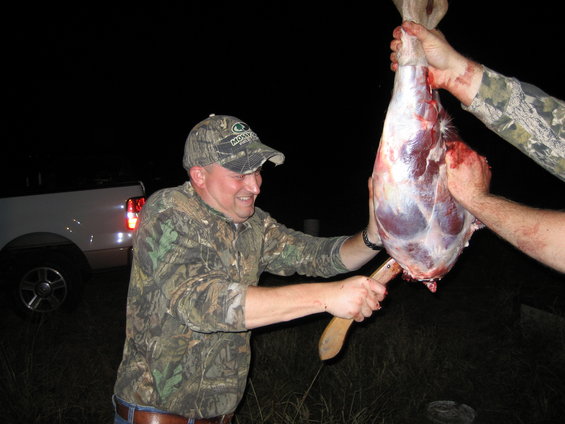 Man in camouflage holding a knife next to a deer carcass being held by two people