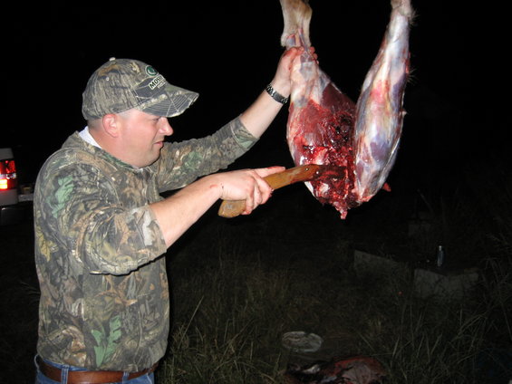 Man in camouflage hat and shirt butchers a deer carcass with a knife at night
