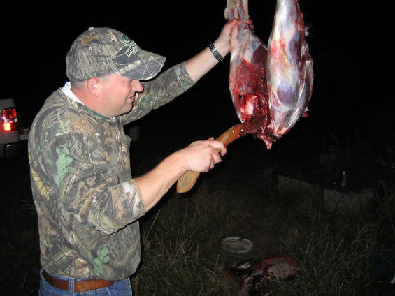 Man cutting hanging animal carcass with knife. Truck taillight visible