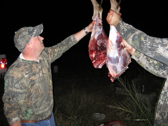 Two men in camouflage clothing holding up freshly butchered deer carcasses outdoors at night