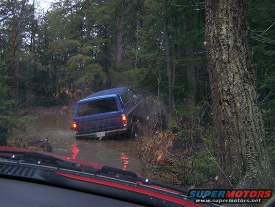 Blue SUV driving through muddy water in a forest. License plate is visible but unreadable