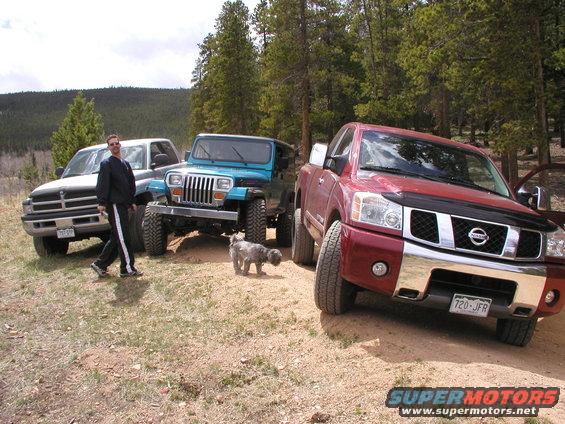 Red Nissan Titan pickup truck parked on dirt road, with a silver Dodge pickup and blue Jeep Wrangler behind it. Man and dog nearby