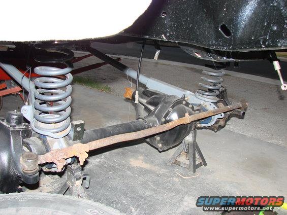 Underside of a vehicle showing the front axle, suspension springs, and steering linkage, all supported by jack stands