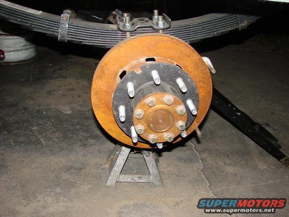 Rusted brake rotor and hub assembly with lug bolts, supported by jack stands. Leaf spring visible above