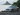 Blurry image of a white car's side profile, possibly a sedan, with dark green foliage in the background