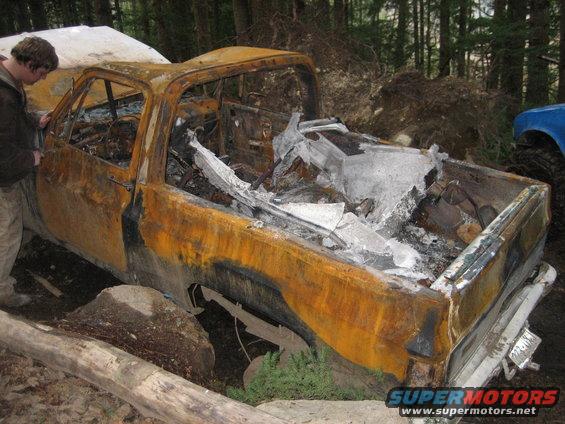 Burnt-out pickup truck with interior destroyed, parked in woods. Blue vehicle visible to the right