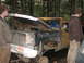 1990 Ford Bronco - This severely damaged pickup truck sits in a wooded area with two onlookers.