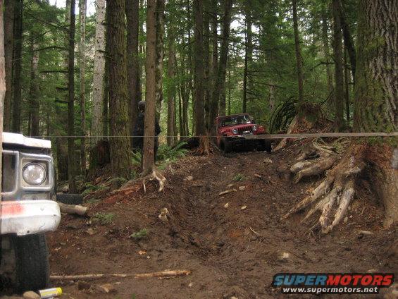 Foreground shows part of a white truck. Background shows a red Jeep on a muddy hill in a forest