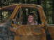 1990 Ford Bronco - A person sits in the driver's seat of a heavily damaged and burnt truck cab, surrounded by trees.