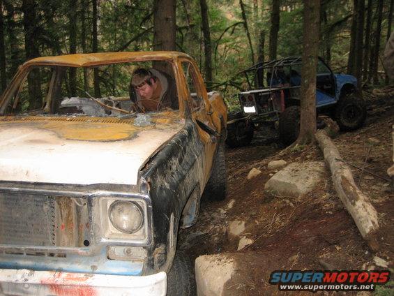 Front view of a wrecked pickup truck with a person inside, and a blue off-road buggy behind it in a forest